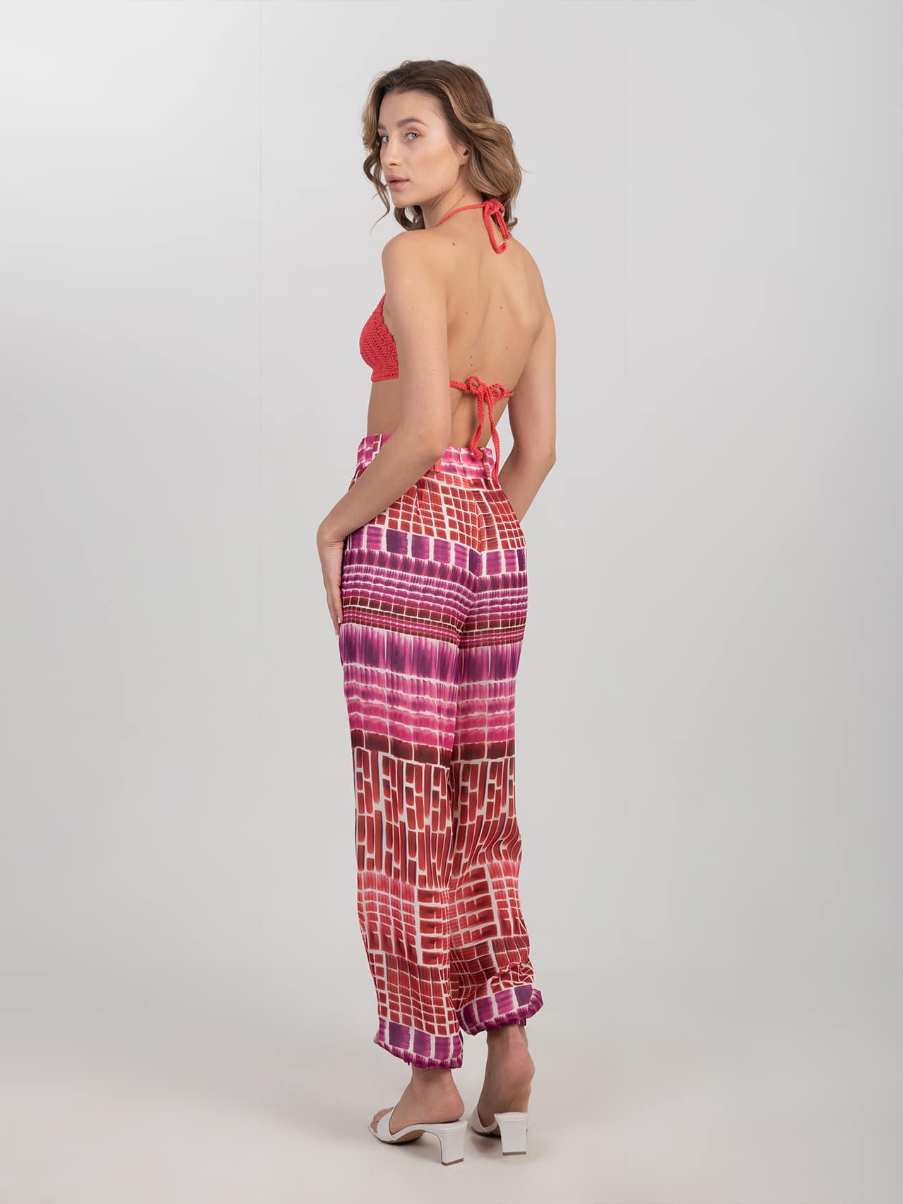 Jedwabne wzorzyste spodnie - LIMITED EDITION Wzorzyste spodnie z jedwabiu w odcieniach czerwieni i różu z wysokim stanem i bufiastymi nogawkami zestawione z czerwonym biustonoszem na szydełku i białymi klapkami na obcasie - widok z tyłu